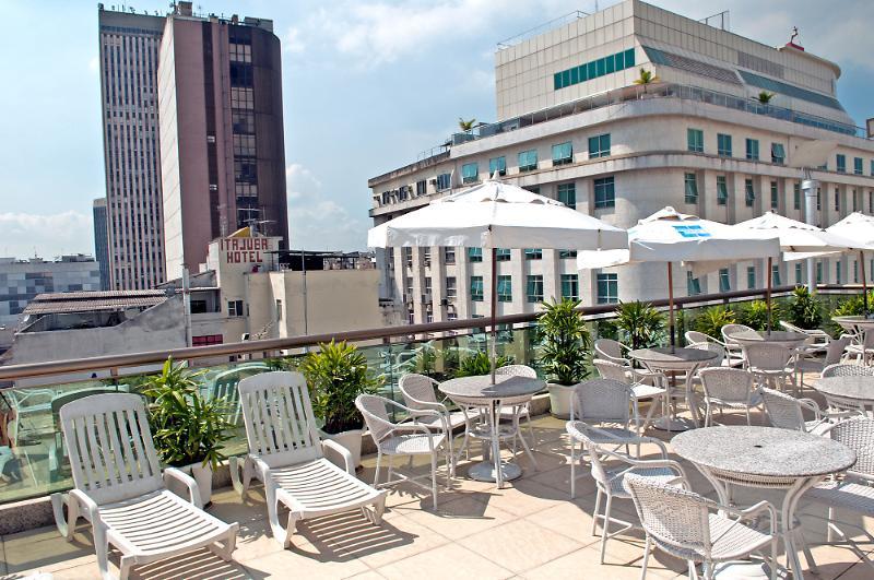 Hotel Atlântico Business Centro Rio de Janeiro Esterno foto The photo shows a rooftop area with several white lounge chairs and circular tables. There are umbrellas providing shade over some of the seating. In the background, there are tall buildings, including a modern skyscraper with a distinct architectura